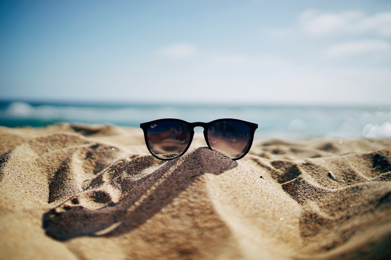 Black sunglasses resting on warm sand with a blurred blue ocean and sky behind
